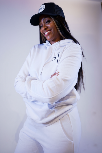 A woman standing and posing in a white hoodie and joggers set, with a cap on her head.